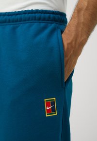 Tyrkysové Nike tepláky majú logo nášivku so žltým prešívaním a červenými, modrými a bielymi akcentmi. Mäkká tkanina s elastickým pásom a bočnou kapsou.