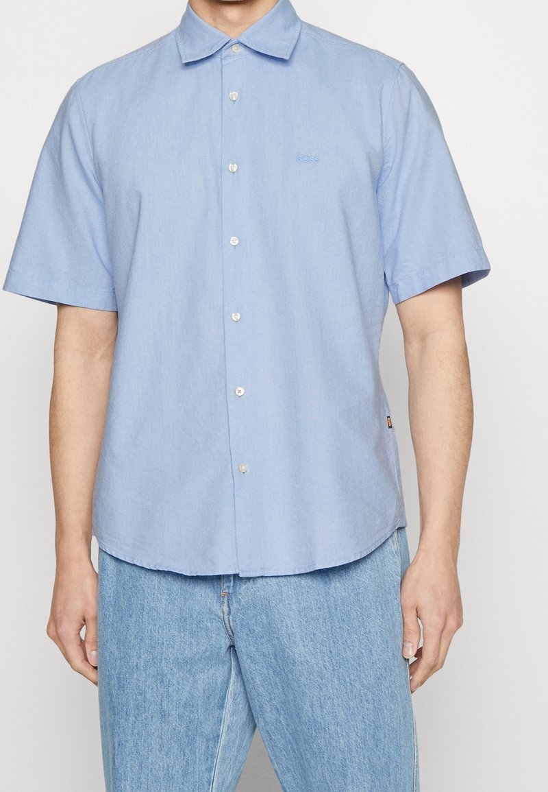 Hombre vestido con una camisa de botones de manga corta azul claro, combinada con unos vaqueros de mezclilla azul claro, de pie contra un fondo liso.