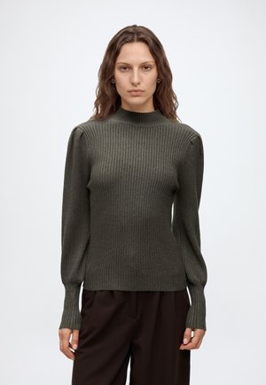 Femme aux cheveux bruns ondulés portant un pull vert olive côtelé à manches longues et un pantalon marron foncé, debout devant un fond uni.