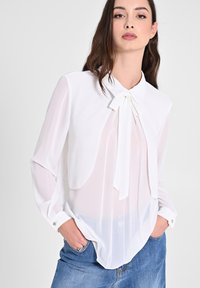 Blusa bianca semitrasparente con colletto a fiocco. Maniche lunghe con polsini abbottonati e una silhouette fluida. Tessuto morbido e leggero.