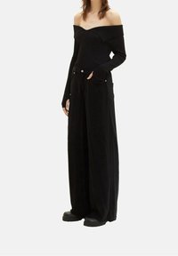 Femme portant un haut noir à manches longues décolleté épaules, pantalon noir à jambes larges, et chaussures à plateforme noires, debout avec une main dans la poche.