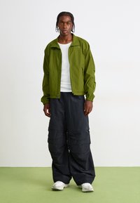 Young man standing on green floor wearing braided hair, green zip-up jacket, white shirt, black wide pants, and white sneakers, facing forward.