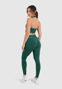 Groene sportbeha en hoge taille legging, beide aansluitend en naadloos, met een matte textuur. Witte branding op de tailleband en de top. Witte sneakers.