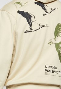 Sweat à capuche beige avec des imprimés d'oies volantes noires et vertes et le texte "UNIFIED PERSPECTIVE" sur la partie inférieure du dos.