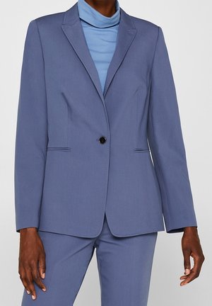 Osoba o ciemnej karnacji ubrana w dopasowaną, niebieską marynarkę i pasujące spodnie, z jasnoniebieskim golfem pod spodem.