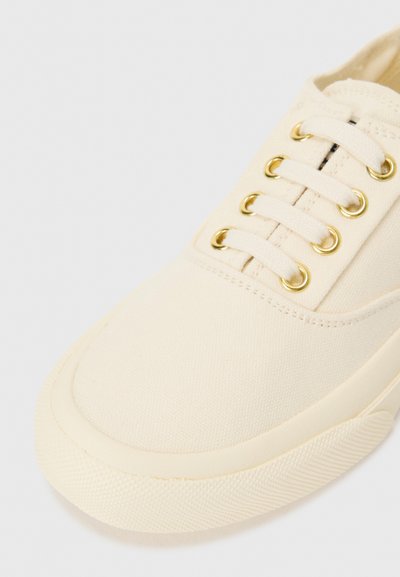 Sneaker in tela color crema con una finitura liscia. Caratterizzato da occhielli dorati e lacci piatti, punta arrotondata e suola in gomma testurizzata per una migliore aderenza.