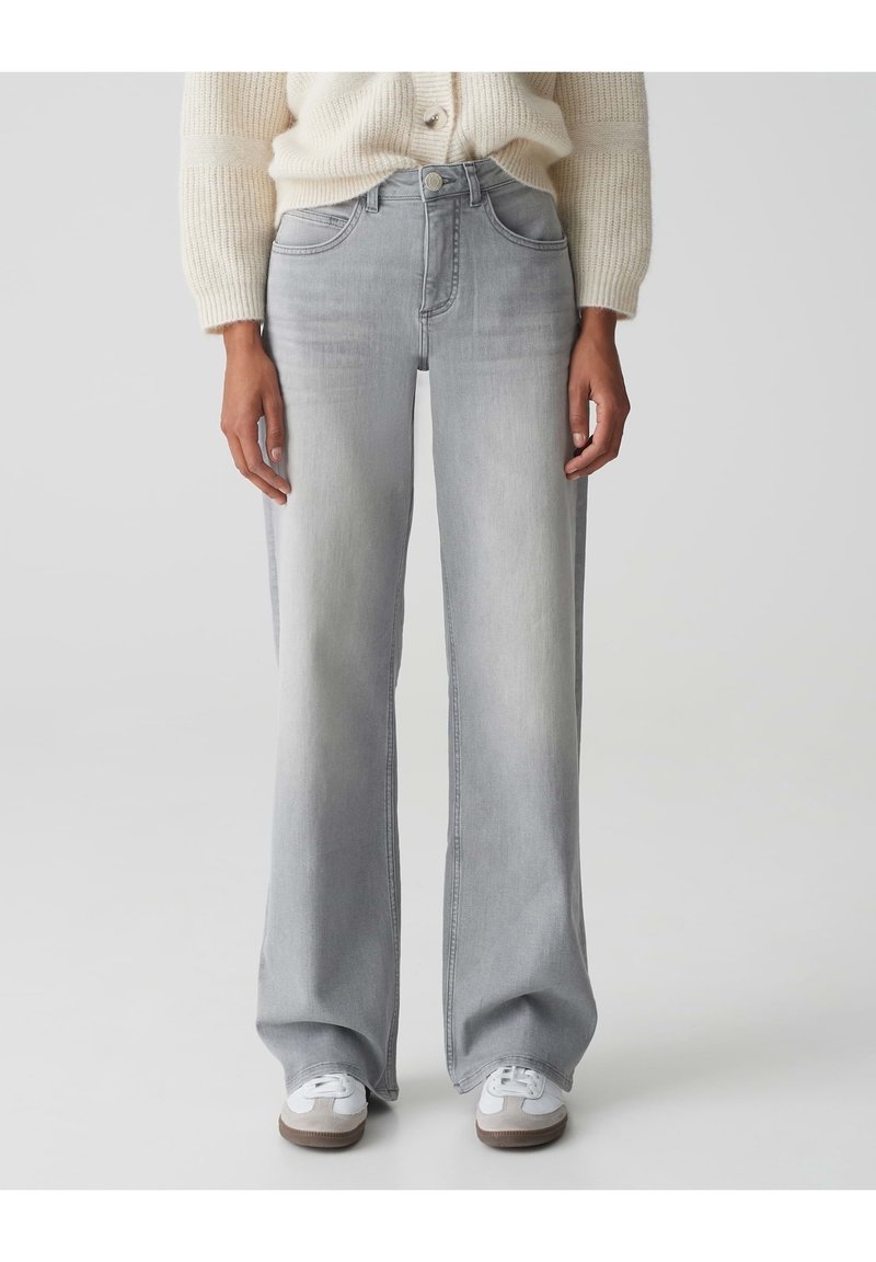 Personne portant un jean large gris clair, un pull en maille crème et des baskets blanches, debout devant un fond uni gris clair.