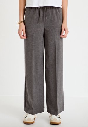 Persoon die grijze broek met wijde pijpen en subtiele krijtstrepen draagt, een wit top, beige en witte sneakers en een armband om de linker pols.