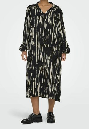 Personne portant une robe boutonnée noire et beige à rayures abstraites, longueur genou, avec de longues manches bouffantes et des chaussures noires brillantes à lacets.