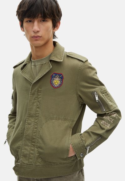 Militaire groene jas met patches op de borst en arm, ritszakken en een gladde, gestructureerde stof.