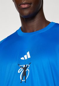 Kék sportpóló sima anyagból, melynek elülső részén fehér grafikával és sziluett mintával díszített. Kerek nyakkivágású fazon.