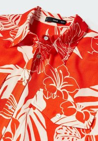 Camicia rossa con un motivo floreale tropicale bianco, maniche corte, colletto e chiusura anteriore con bottoni. Presenta due tasche frontali con chiusure a bottone.