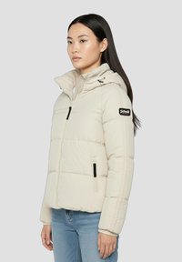 Beige dunjakke med hætte, der har lynlås, sidelommer og et logopatch på ærmet. Glat tekstur og quiltet design.