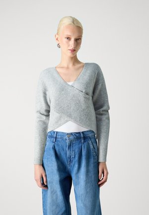 Maglione grigio chiaro, stile avvolgente con scollatura a V e maniche lunghe. Indossato sopra una maglietta bianca, abbinato a jeans di denim blu a vita alta.