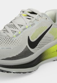 Крупен план на бяла маратонка с черно лого на Nike, светло зелени и жълти акценти, дишаща мрежеста горна част и бели връзки на сив фон.