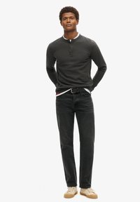 Camisa henley negra de manga larga y canalé con tapeta de botones, combinada con jeans oscuros y zapatillas beige. Diseño minimalista con acentos en contraste.