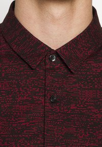 Gros plan sur une personne portant une chemise noire à motif de points rouges, boutonnée au col et sur la partie supérieure de la poitrine.
