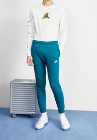 Vit långärmad skjorta med en flerfärgad grafik på bröstet, i kombination med teal färgade mjukisbyxor och vita sneakers. Texturerade tygdetaljer synliga.