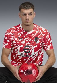 Raudona maskuojanti sportinė marškinė su juodais akcentais ir logotipu; raudonas futbolas su juodais detalėmis, laikomas rankose.