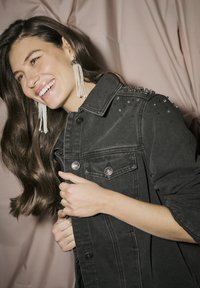 Jachetă din denim negru cu embellishments cu tinte pe umeri, nasturi frontali și două buzunare. Modelul poartă cercei lungi cu perle, zâmbind.