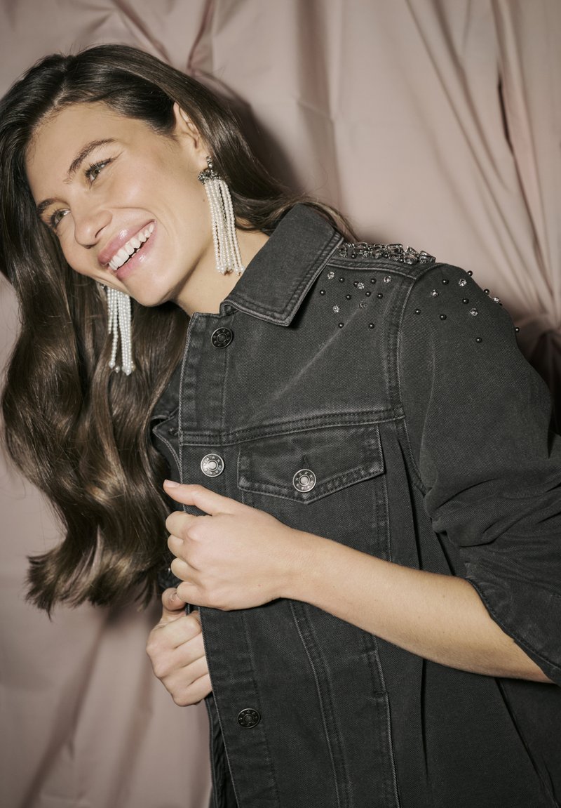Jachetă din denim negru cu embellishments cu tinte pe umeri, nasturi frontali și două buzunare. Modelul poartă cercei lungi cu perle, zâmbind.