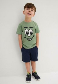 T-shirt in cotone verde con un grande design di faccia da cartone animato, abbinata a pantaloni corte blu navy e sneakers nere con accenti verdi.
