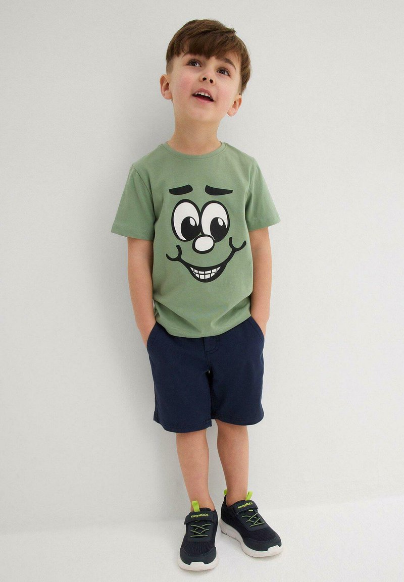 T-shirt in cotone verde con un grande design di faccia da cartone animato, abbinata a pantaloni corte blu navy e sneakers nere con accenti verdi.