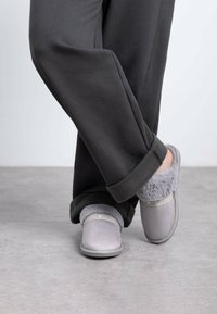 Zapatillas gris con un suave forro de piel, con una suela plana y una banda de acento sutil en la parte superior. Llevadas con pantalones oscuros de corte holgado.