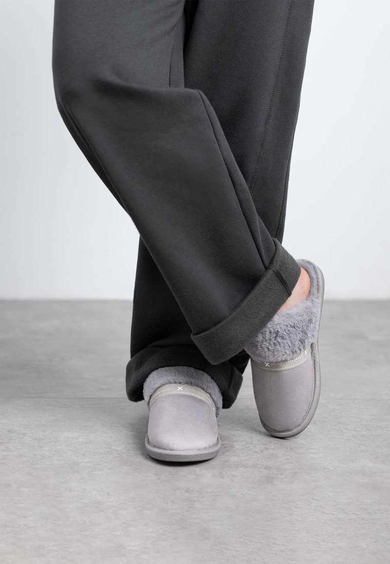 Zapatillas gris con un suave forro de piel, con una suela plana y una banda de acento sutil en la parte superior. Llevadas con pantalones oscuros de corte holgado.