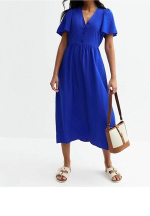 Frau trägt ein blaues Midikleid mit flatternden Ärmeln, weiße Sandalen mit Schnalle und hält eine cremefarbene und braune Handtasche am Trageriemen.