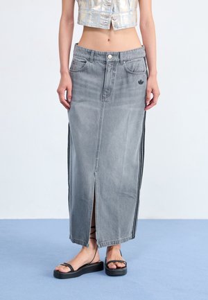 Donna che indossa una lunga gonna di jeans grigia con spacco frontale, abbinata a sandali platform neri e un gilet metallico corto, in piedi su pavimento blu.