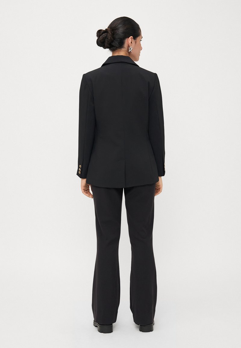 Schwarzer taillierter Blazer und ausgestellte Hose. Der Blazer hat einen Reverskragen und goldene Knopfakzente. Glattes, strukturiertes Material.