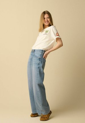 Jeune femme souriante, portant un t-shirt blanc à motifs, un jean bleu clair ample et des mocassins marron, debout devant un fond beige.