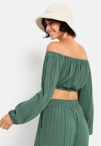 Πράσινη off-the-shoulder crop top με μακριά μανίκια, υφασμάτινη υφή και ελαστικό τελείωμα, συνδυασμένο με ψηλόμεσα παντελόνια και έναν κρεμ καπέλο bucket.