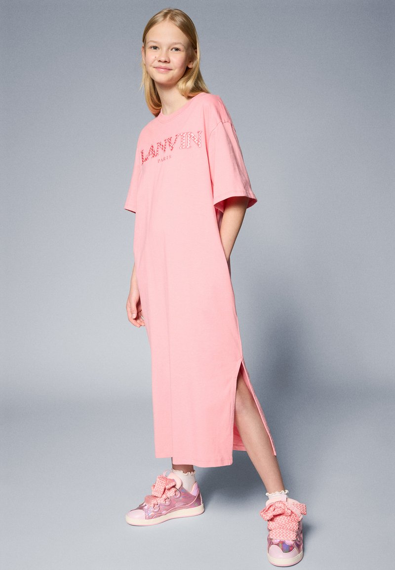 Vestido rosa oversize con mangas cortas, que presenta el nombre de la marca "LANVIN" en una tipografía decorativa; tiene aberturas laterales y se lleva con zapatillas adornadas.