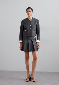 Grijze geruite cropped jacket met gouden knopen, gecombineerd met een geplooide rok met zijaccenten. Witte manchetten op de jacket. Zwarte sandaaltjes met bandjes.
