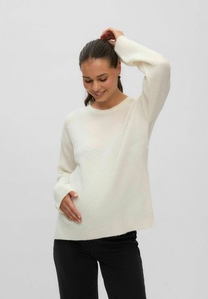 Femme souriante, portant un pull blanc et un pantalon noir, une main dans le dos et l'autre touchant ses cheveux sur un fond uni.