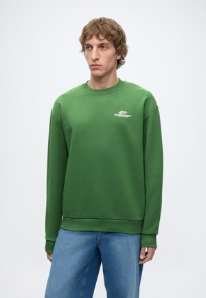 Jeune homme aux cheveux bouclés châtain clair portant un sweat-shirt vert à col rond avec un petit logo blanc et un jean bleu ample sur fond blanc.