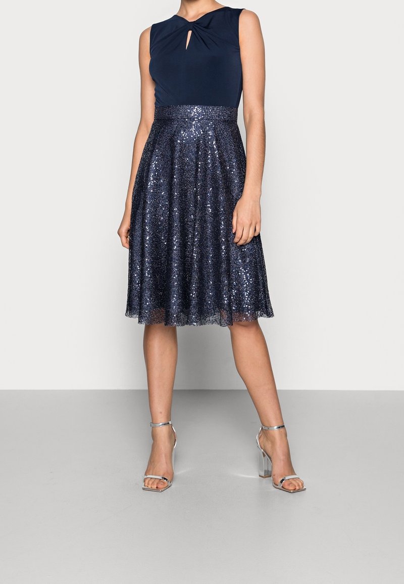 Femme portant un haut sans manches bleu marine avec un détail œillet et une jupe bleu marine à sequins longueur genou avec des sandales argentées à talons hauts.