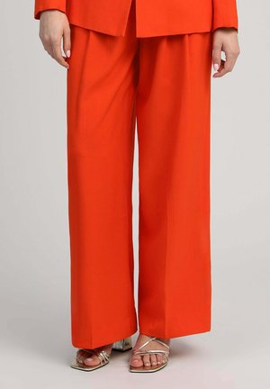 Femme portant un pantalon large orange vif et des sandales argentées métalliques à bout ouvert, debout devant un fond neutre.