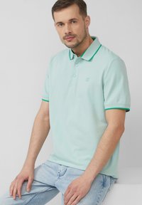 Polo shirt verde chiaro con colletto, bordi verdi a contrasto e chiusura con tre bottoni; abbinato a jeans azzurri.