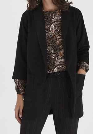 Kvinde iført en sort åben blazer over en paisley-mønstret langærmet top og mørke ternede bukser med snøre i taljen.