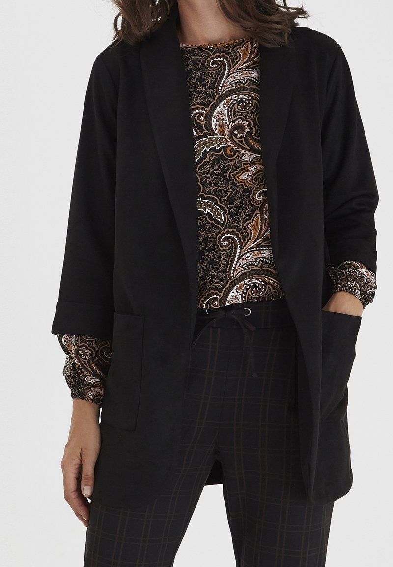 Femme portant un blazer noir ouvert sur un top à manches longues à motif paisley et un pantalon à carreaux foncé avec une taille à cordon.