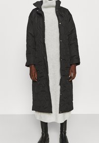 Cappotto lungo trapuntato nero con colletto alto, chiusure a scatto e tessuto testurizzato, indossato sopra un vestito in maglia grigio chiaro e stivali neri.