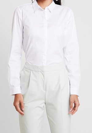 Femme portant une chemise blanche cintrée boutonnée rentrée dans un pantalon gris clair taille haute sur un fond uni.