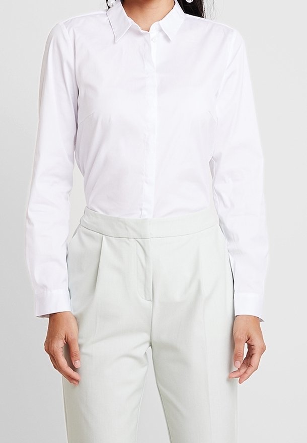 Femme portant une chemise blanche cintrée boutonnée rentrée dans un pantalon gris clair taille haute sur un fond uni.