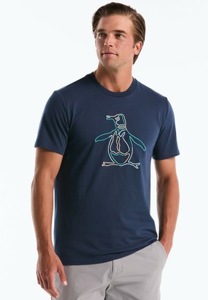 Mann trägt dunkelblaue T-Shirt mit buntem Neon-Pinguin-Design, steht mit einer Hand in der Tasche vor hellem Hintergrund.