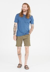 Blårandig t-shirt, khaki-shorts och bruna sandaler. T-shirten har rund halsringning; shortsen är upprullade vid fållen. Tatueringar synliga på benen.