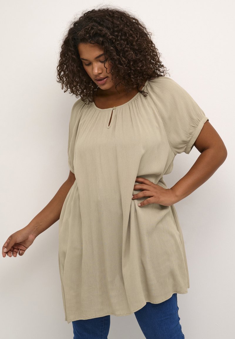 Tunika di un beige chiaro con vestibilità ampia, scollatura arricciata e maniche corte. Presenta un dettaglio a chiave e un tessuto morbido e testurizzato. Abbinata a jeans blu.