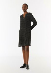 Schwarzes langärmliges Kleid mit V-Ausschnitt, das ein strukturiertes Muster und einen plissierten Rock aufweist. Knie-lange Länge mit seitlichen Taschen. Getragen mit schwarzen Schuhen.